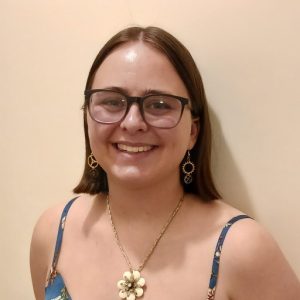 A photo of Linnea Peterson, a white femme-presenting person with brown shoulder-length hair, wearing gray glasses, steampunk earrings, a flower necklace, and a strappy dress